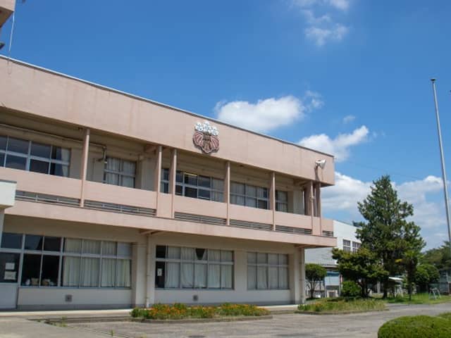 青空の下に建つ日本の公立学校の校舎、校章と整った花壇が見える。