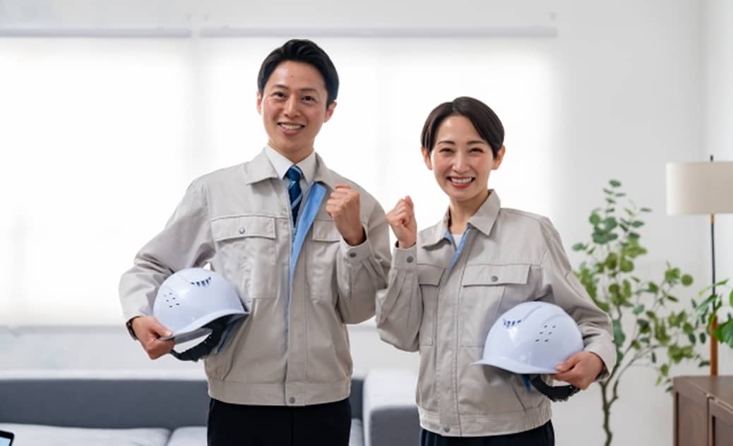 作業服を着た男女がヘルメットを手に持ち、笑顔でガッツポーズをしている写真