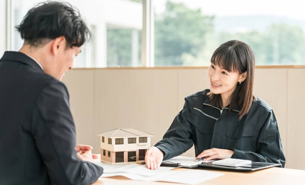 作業服姿の女性が住宅模型を前に、男性に図面を説明している場面